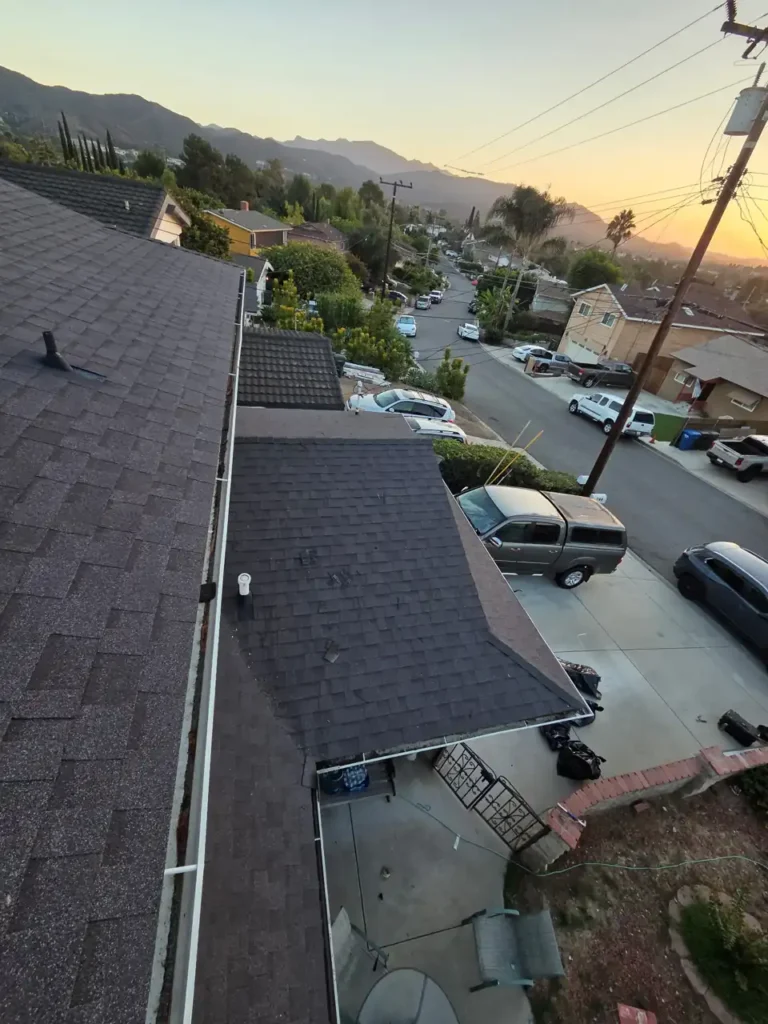 Dark gray shingle roof sections along a gutter on a hillside street at sunset, with mountains in the distance