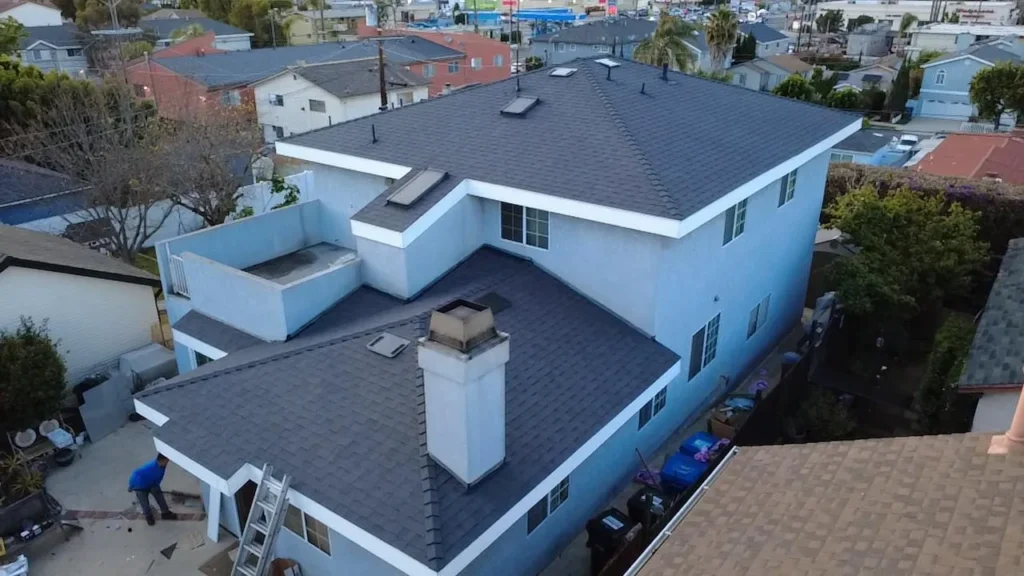 Aerial view of a two-story house with a finished gray shingle roof, skylights and a stucco chimney, a crew member and ladder at the side