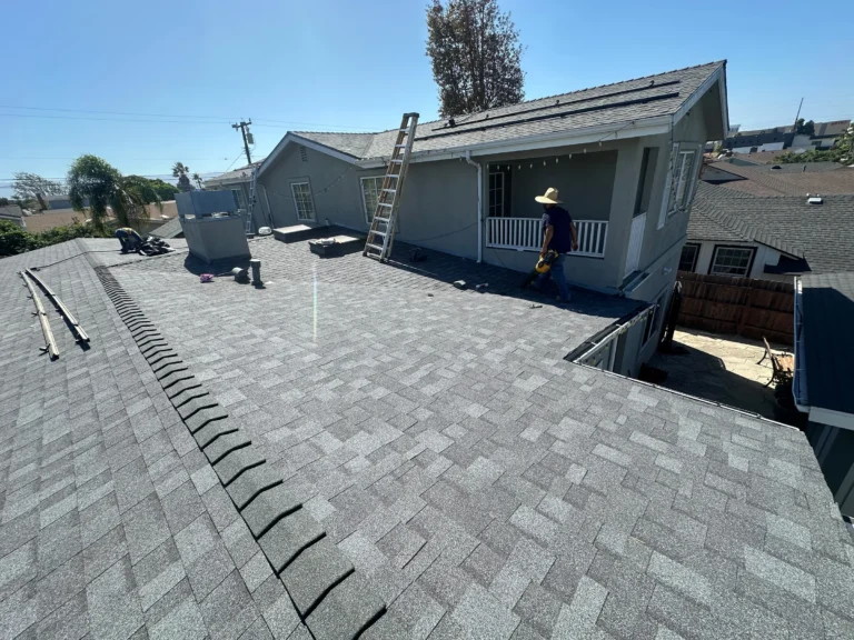 New gray shingle roof with a ridge cap, a ladder and a worker on the porch roof, solar panels in the background