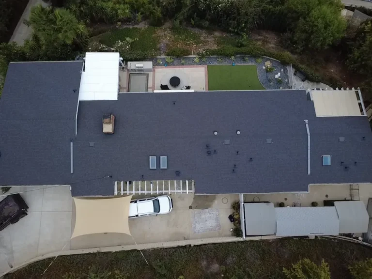 Top-down aerial view of a long single-story house with a new dark gray shingle roof, skylights and a patio with shade sails