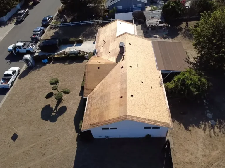 Aerial view of a single-story house with the old roof removed and new plywood decking installed, work trucks parked at the curb
