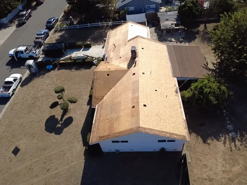 Aerial view of a single-story house with the old roof removed and new plywood decking installed, work trucks parked at the curb
